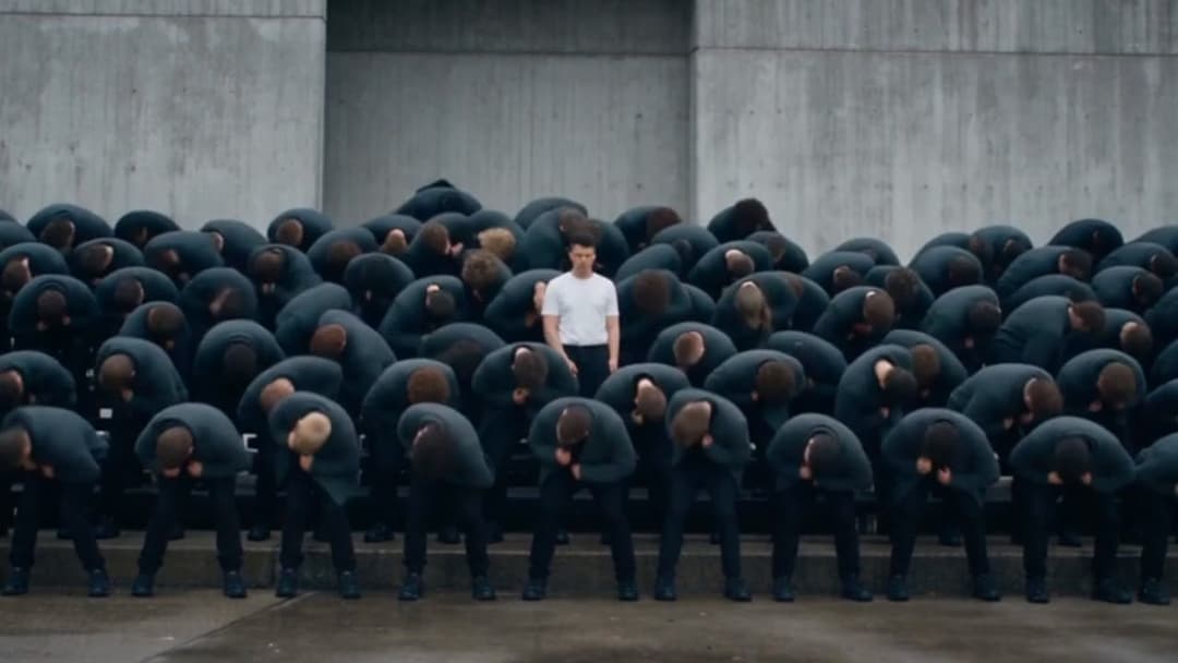 Eighty figures in charcoal hoodies bow in unison on concrete steps; one man in a white t-shirt stands upright in the middle of them