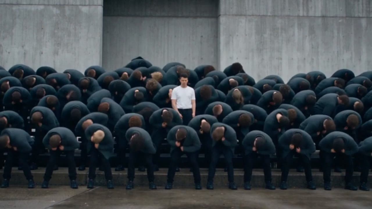 Eighty figures in charcoal hoodies bow in unison on concrete steps; one man in a white t-shirt stands upright in the middle of them